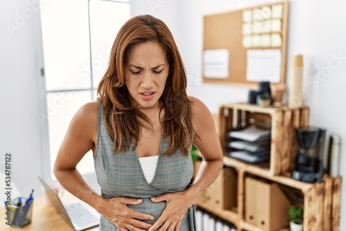 Middle age hispanic woman at the office with hand on stomach because indigestion, painful illness feeling unwell. ache concept.