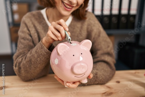 Young caucasian woman ecommerce business worker insert dollar on piggy bank at office