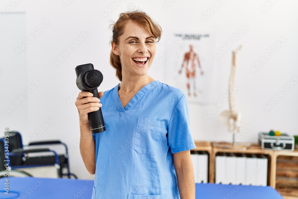 Young caucasian physio therapist girl smiling happy holding gun percusion at the clinic.