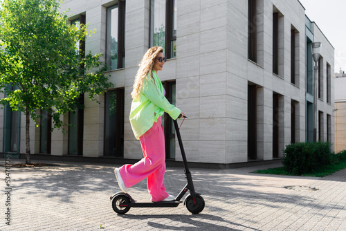 Young Beautiful Woman Dressed Pink Jeans And Bright Lime Colored Shirt And White Sneakers Riding Black Electric Scooter photo