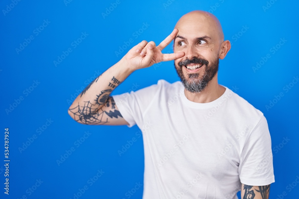 Naklejka premium Hispanic man with tattoos standing over blue background doing peace symbol with fingers over face, smiling cheerful showing victory
