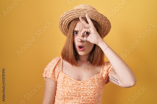 Young redhead woman standing over yellow background wearing summer hat doing ok gesture shocked with surprised face, eye looking through fingers. unbelieving expression.