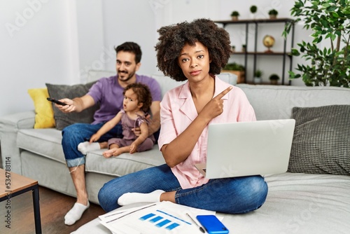 Mother of interracial family working using computer laptop at home pointing with hand finger to the side showing advertisement, serious and calm face