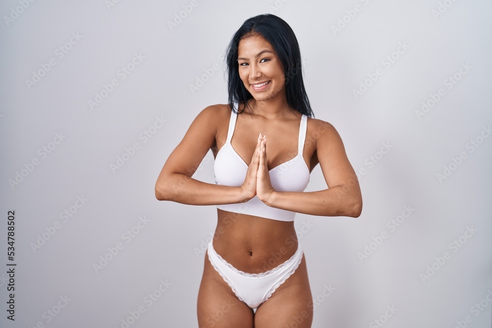 Hispanic woman wearing lingerie praying with hands together asking for forgiveness smiling confident. Hispanic woman wearing lingerie praying with hands together asking for forgiveness smiling confident.