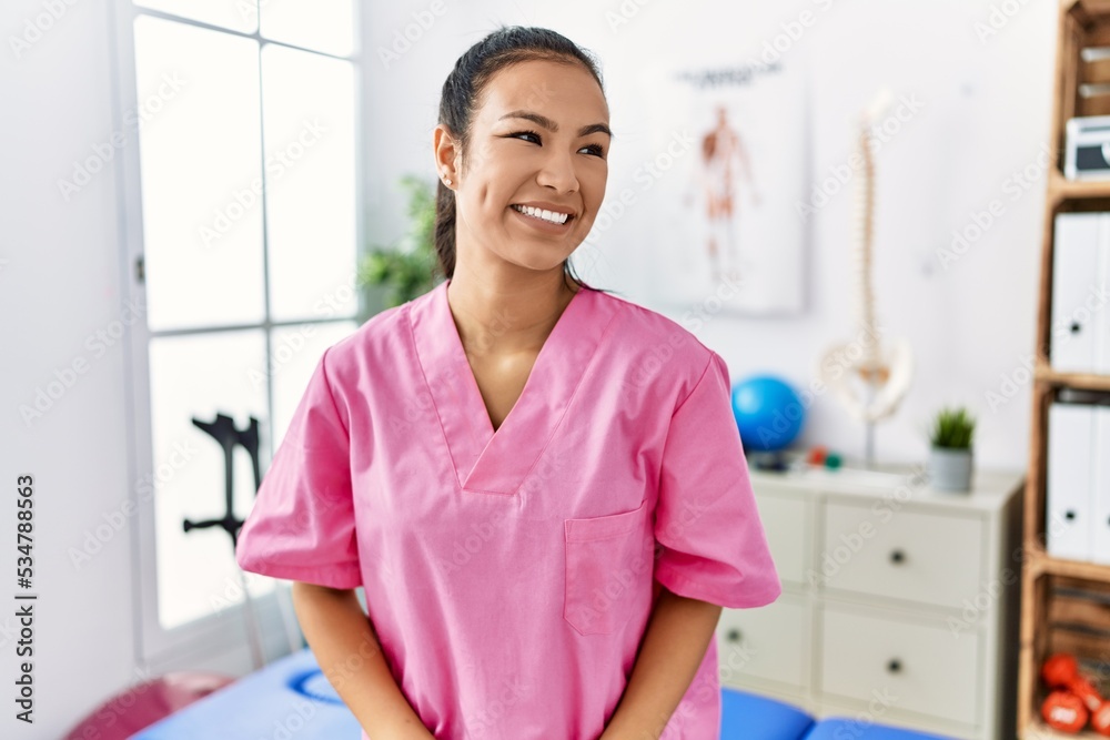 Young hispanic woman working at pain recovery clinic looking away to side with smile on face, natural expression. laughing confident. Young hispanic woman working at pain recovery clinic looking away to side with smile on face, natural expression. laughing confident.