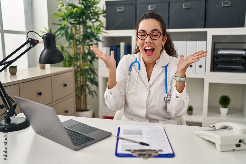 Young hispanic woman wearing doctor uniform and stethoscope celebrating mad and crazy for success with arms raised and closed eyes screaming excited. winner concept Young hispanic woman wearing doctor uniform and stethoscope celebrating mad and crazy for success with arms raised and closed eyes screaming excited. winner concept