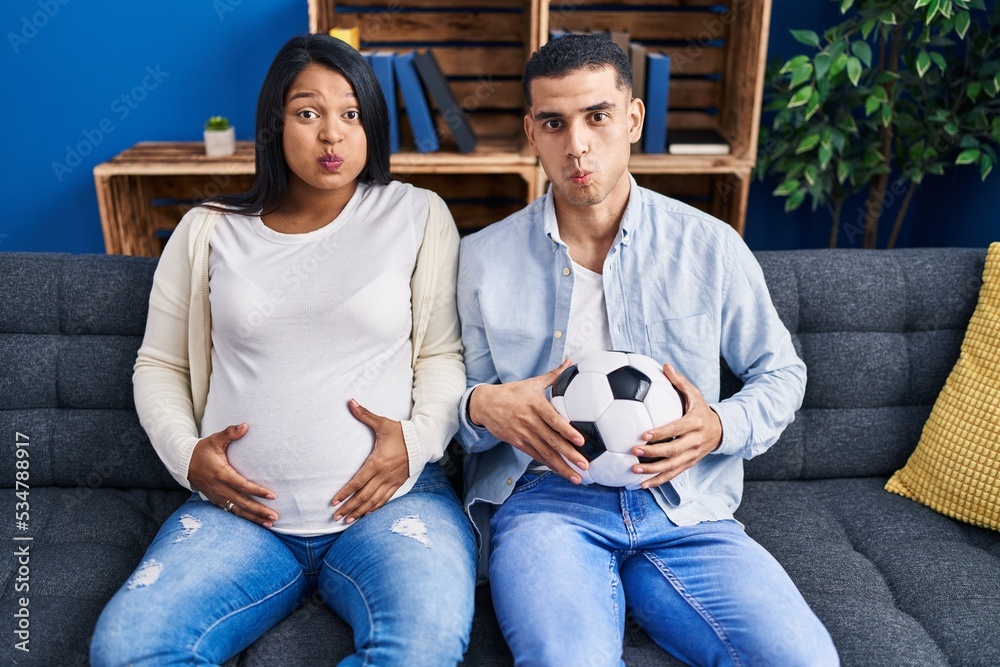Young hispanic couple expecting a baby sitting on the sofa holding ball and tummy puffing cheeks with funny face. mouth inflated with air, catching air. - obrazy, fototapety, plakaty 