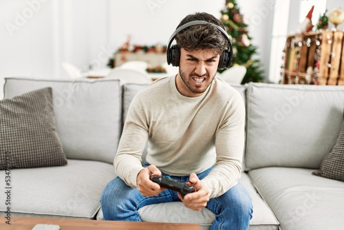 Young hispanic man angry sitting on the sofa playing video game at home.