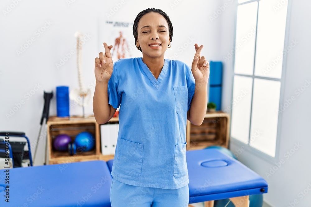 Young african american woman working at pain recovery clinic gesturing finger crossed smiling with hope and eyes closed. luck and superstitious concept. Young african american woman working at pain recovery clinic gesturing finger crossed smiling with hope and eyes closed. luck and superstitious concept.