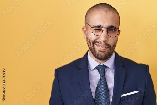 Hispanic man with beard wearing suit and tie winking looking at the camera with sexy expression, cheerful and happy face.