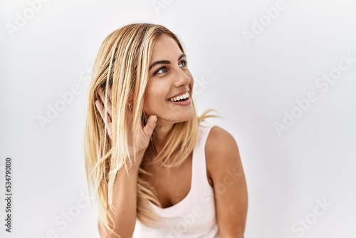 Blonde beautiful young woman standing over white isolated background smiling with hand over ear listening an hearing to rumor or gossip. deafness concept.