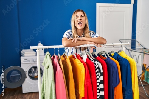 Young blonde woman at laundry room with clean clothes angry and mad screaming frustrated and furious, shouting with anger looking up.