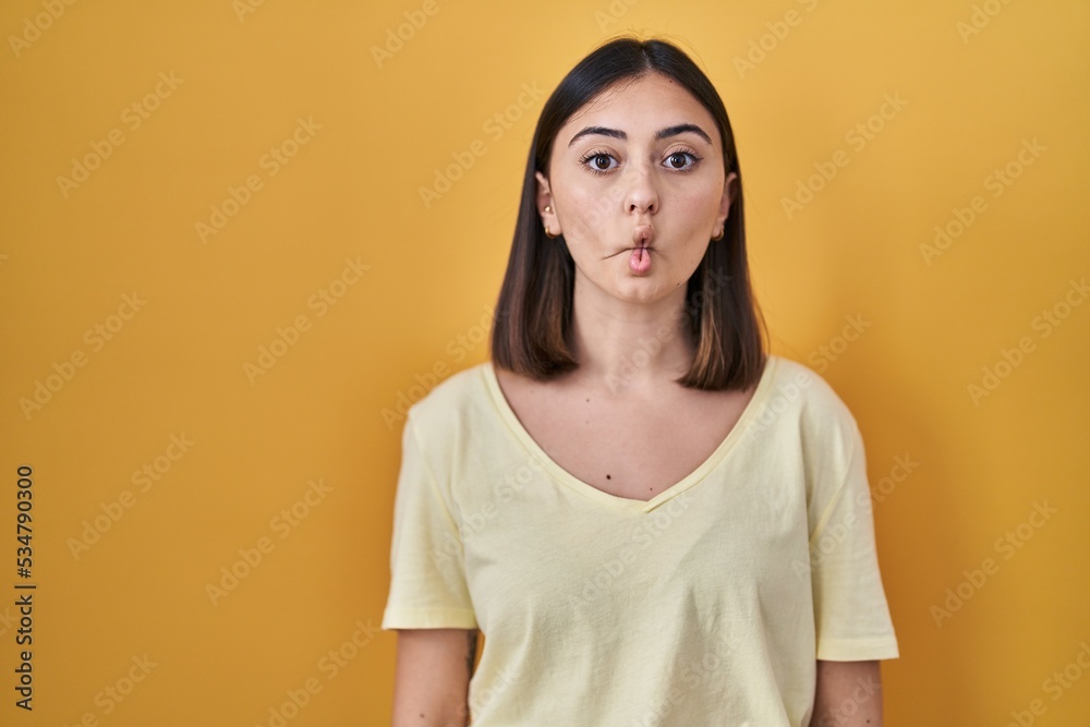 Hispanic girl wearing casual t shirt over yellow background making fish face with lips, crazy and comical gesture. funny expression.