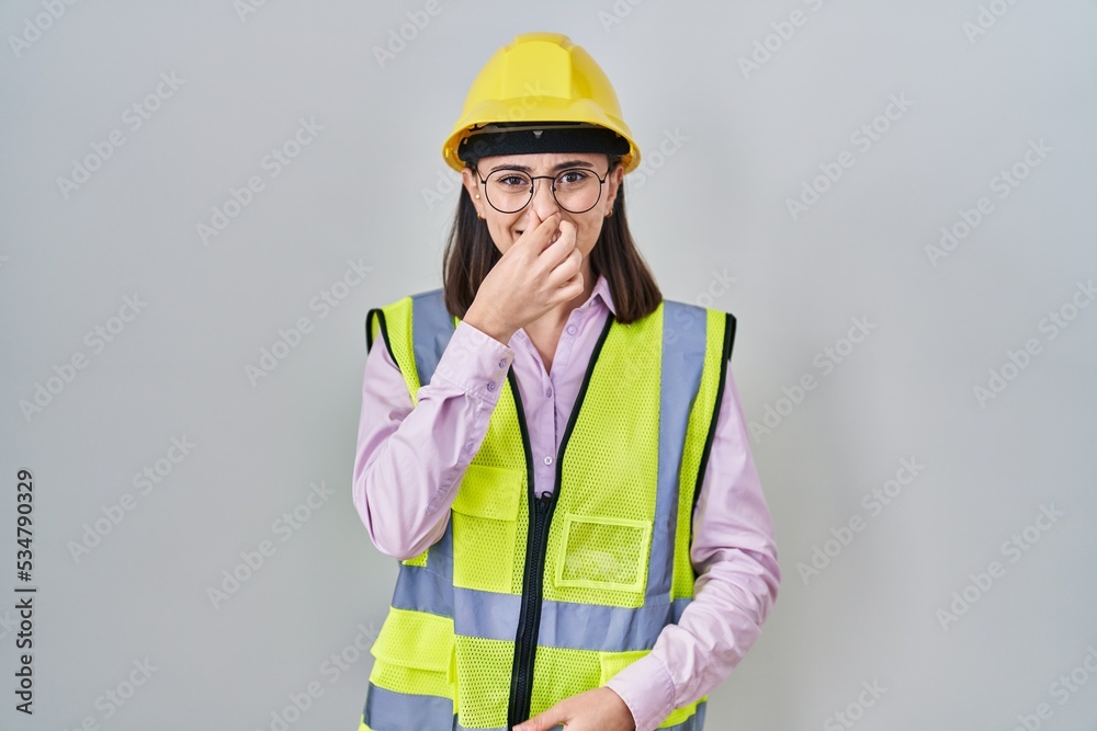 Hispanic girl wearing builder uniform and hardhat smelling something stinky and disgusting, intolerable smell, holding breath with fingers on nose. bad smell Hispanic girl wearing builder uniform and hardhat smelling something stinky and disgusting, intolerable smell, holding breath with fingers on nose. bad smell