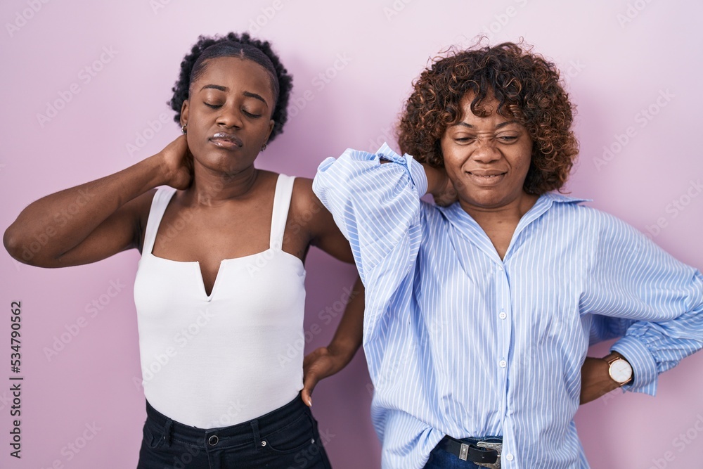 Two african women standing over pink background suffering of neck ache injury, touching neck with hand, muscular pain Two african women standing over pink background suffering of neck ache injury, touching neck with hand, muscular pain