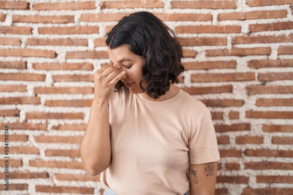 Young hispanic woman standing over bricks wall tired rubbing nose and eyes feeling fatigue and headache. stress and frustration concept. Young hispanic woman standing over bricks wall tired rubbing nose and eyes feeling fatigue and headache. stress and frustration concept.