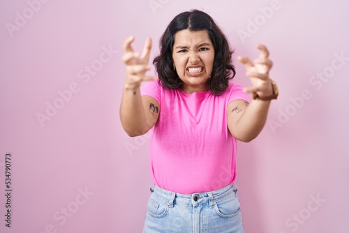 Young hispanic woman standing over pink background shouting frustrated with rage  hands trying to strangle  yelling mad
