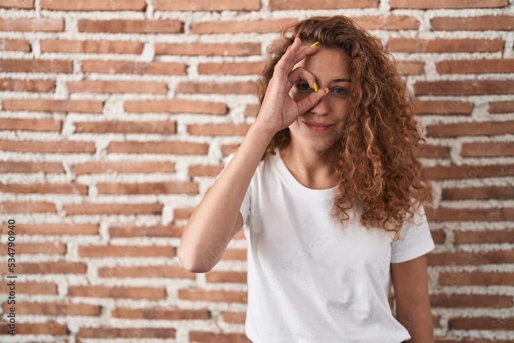 Young caucasian woman standing over bricks wall background doing ok gesture with hand smiling, eye looking through fingers with happy face. Young caucasian woman standing over bricks wall background doing ok gesture with hand smiling, eye looking through fingers with happy face.