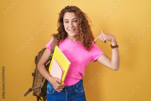 Young caucasian woman wearing student backpack and holding books looking confident with smile on face  pointing oneself with fingers proud and happy.