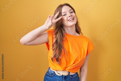 Caucasian woman standing over yellow background doing peace symbol with fingers over face, smiling cheerful showing victory