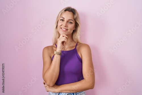 Young blonde woman standing over pink background looking confident at the camera smiling with crossed arms and hand raised on chin. thinking positive.