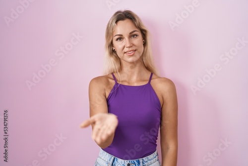 Young blonde woman standing over pink background smiling cheerful offering palm hand giving assistance and acceptance.
