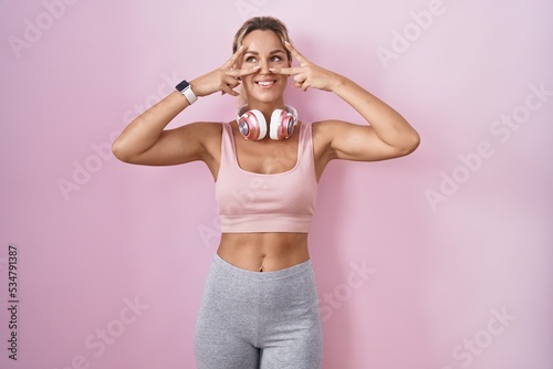 Young blonde woman wearing sportswear and headphones doing peace symbol with fingers over face  smiling cheerful showing victory
