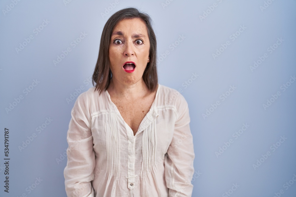 Middle age hispanic woman standing over blue background in shock face, looking skeptical and sarcastic, surprised with open mouth Middle age hispanic woman standing over blue background in shock face, looking skeptical and sarcastic, surprised with open mouth