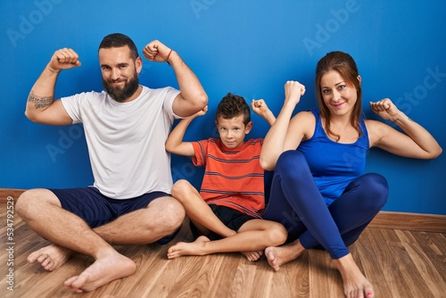 Family of three sitting on the floor at home showing arms muscles smiling proud. fitness concept.