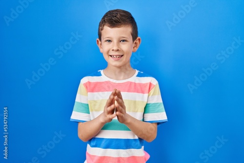 Young caucasian kid standing over blue background hands together and fingers crossed smiling relaxed and cheerful. success and optimistic