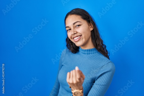 Young brazilian woman standing over blue isolated background beckoning come here gesture with hand inviting welcoming happy and smiling