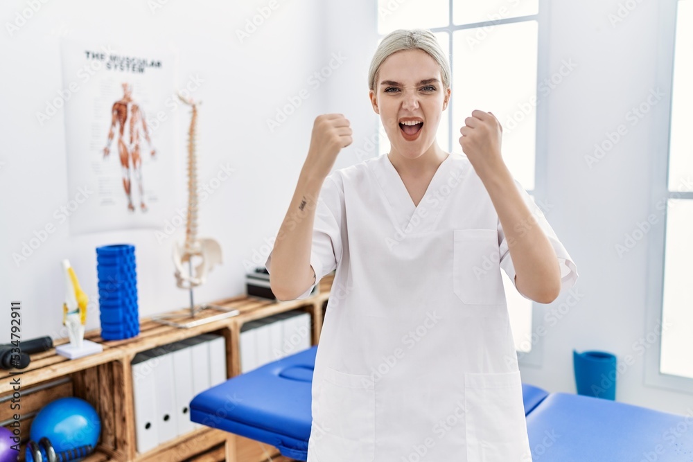 Obraz premium Young caucasian woman working at pain recovery clinic angry and mad raising fists frustrated and furious while shouting with anger. rage and aggressive concept.