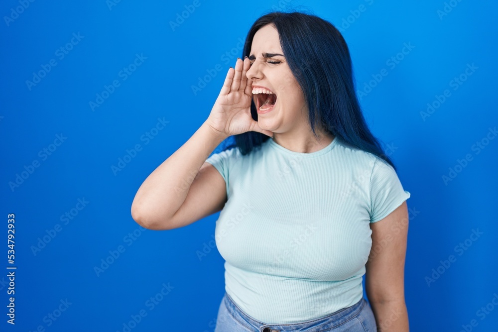 Young modern girl with blue hair standing over blue background shouting and screaming loud to side with hand on mouth. communication concept. Young modern girl with blue hair standing over blue background shouting and screaming loud to side with hand on mouth. communication concept.