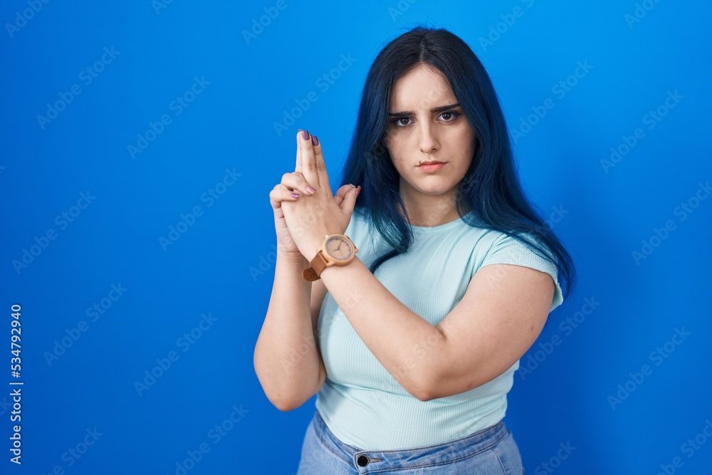 Young modern girl with blue hair standing over blue background holding symbolic gun with hand gesture, playing killing shooting weapons, angry face Young modern girl with blue hair standing over blue background holding symbolic gun with hand gesture, playing killing shooting weapons, angry face