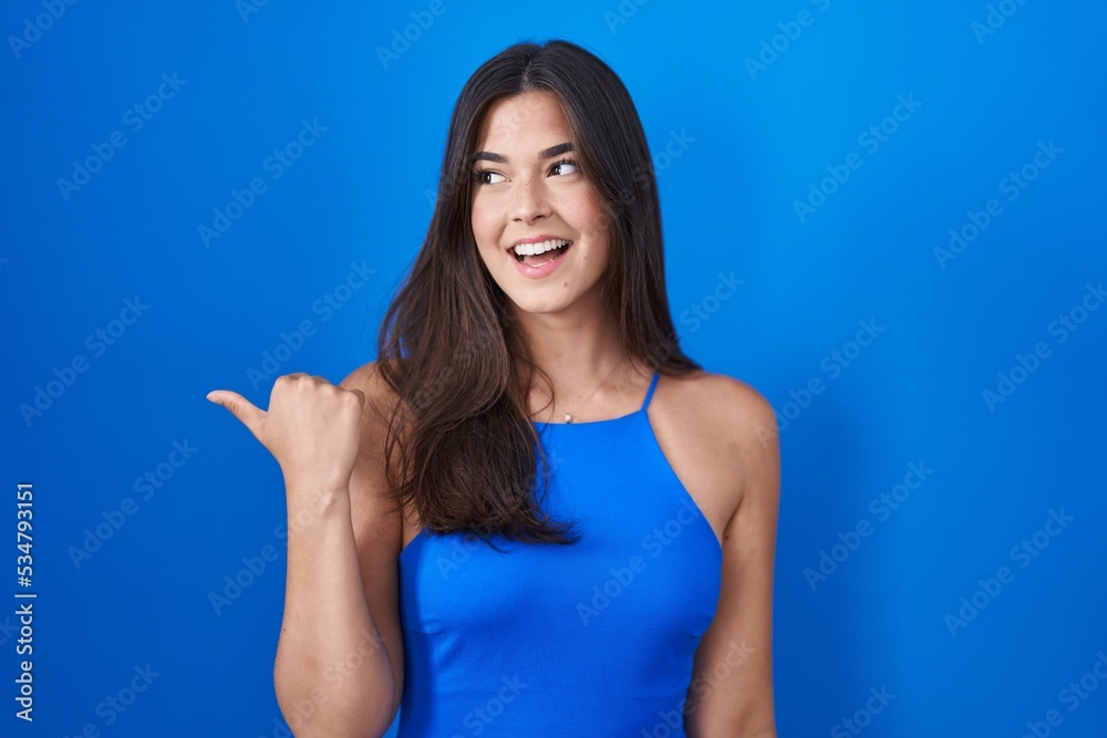 Hispanic woman standing over blue background smiling with happy face looking and pointing to the side with thumb up. Hispanic woman standing over blue background smiling with happy face looking and pointing to the side with thumb up.