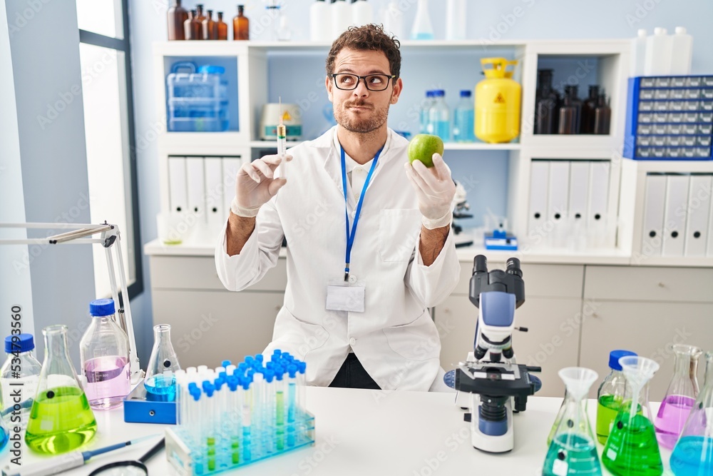 Young hispanic man working at scientist laboratory holding apple smiling looking to the side and staring away thinking. Young hispanic man working at scientist laboratory holding apple smiling looking to the side and staring away thinking.