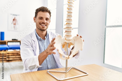 Young hispanic man wearing physiotherapist uniform touching anatomical model of vertebral column at clinic