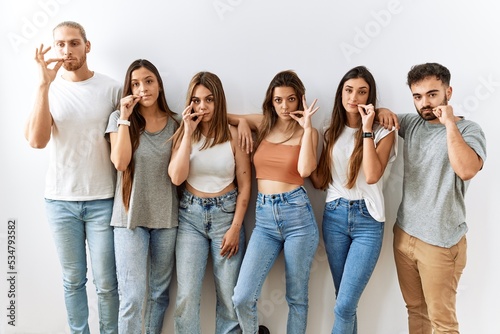 Group of young friends standing together over isolated background mouth and lips shut as zip with fingers. secret and silent, taboo talking photo