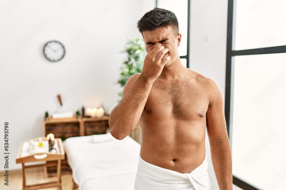 Obraz premium Young hispanic man standing shirtless at spa center smelling something stinky and disgusting, intolerable smell, holding breath with fingers on nose. bad smell