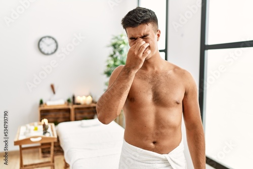 Young hispanic man standing shirtless at spa center smelling something stinky and disgusting, intolerable smell, holding breath with fingers on nose. bad smell