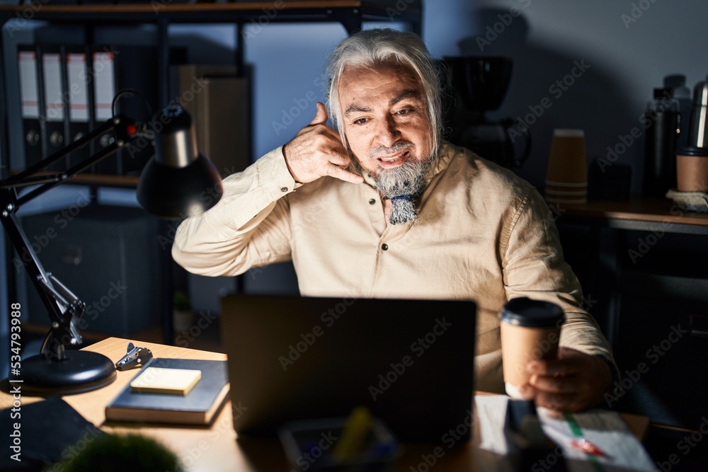 Middle age man with grey hair working at the office at night smiling doing phone gesture with hand and fingers like talking on the telephone. communicating concepts. Middle age man with grey hair working at the office at night smiling doing phone gesture with hand and fingers like talking on the telephone. communicating concepts.
