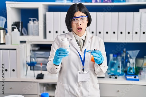 Young asian woman working at scientist laboratory in shock face, looking skeptical and sarcastic, surprised with open mouth