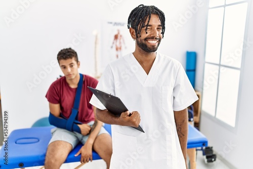 Young hispanic man working at pain recovery clinic with a man with broken arm winking looking at the camera with sexy expression, cheerful and happy face.