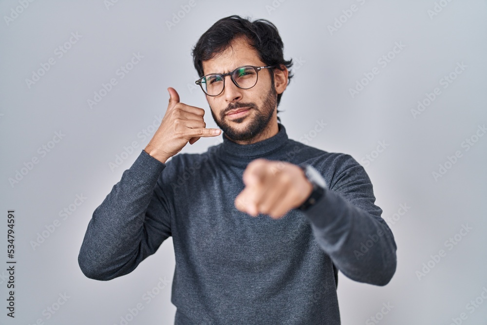 Fototapeta premium Handsome latin man standing over isolated background smiling doing talking on the telephone gesture and pointing to you. call me.