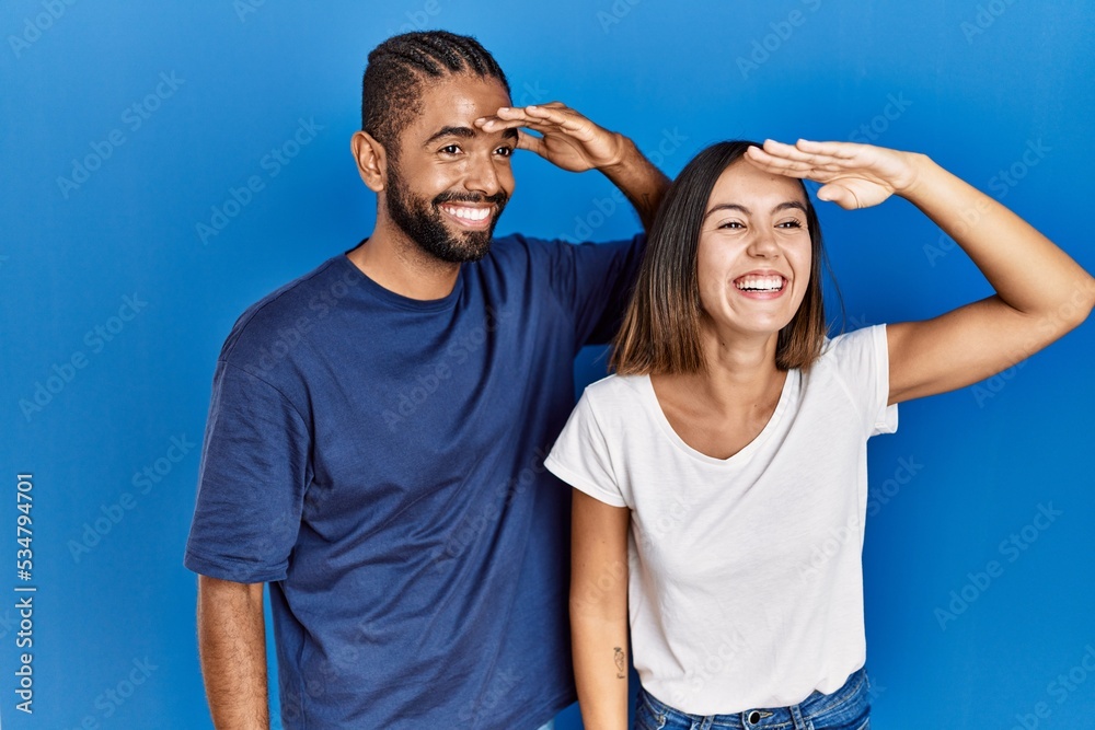 Young hispanic couple standing together very happy and smiling looking far away with hand over head. searching concept. Young hispanic couple standing together very happy and smiling looking far away with hand over head. searching concept.