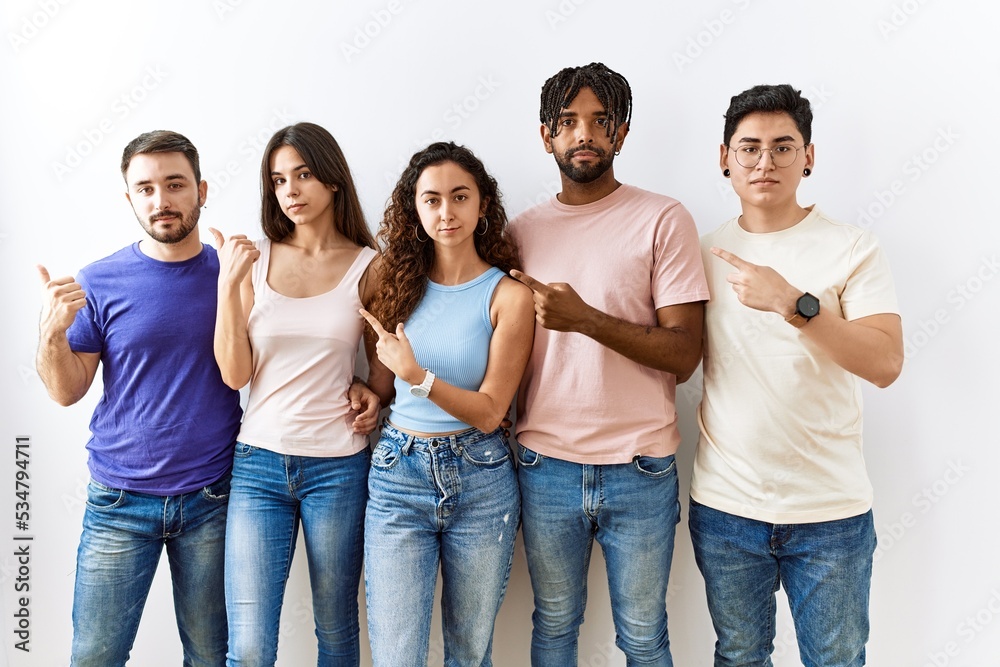 Group of young people standing together over isolated background pointing with hand finger to the side showing advertisement, serious and calm face Group of young people standing together over isolated background pointing with hand finger to the side showing advertisement, serious and calm face