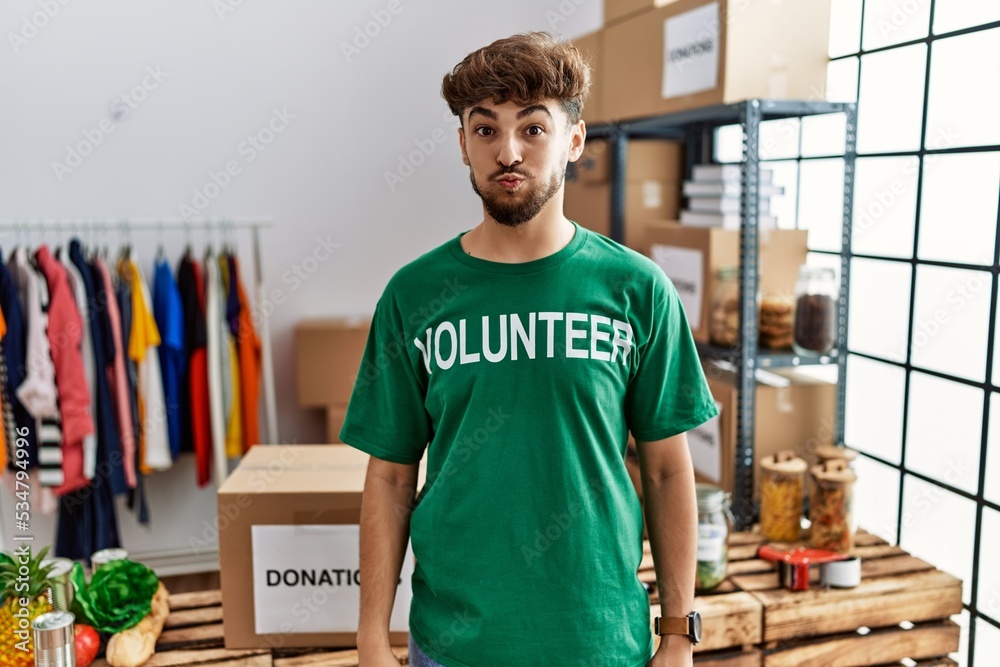 Young arab man wearing volunteer t shirt at donations stand puffing cheeks with funny face. mouth inflated with air, crazy expression. - obrazy, fototapety, plakaty 