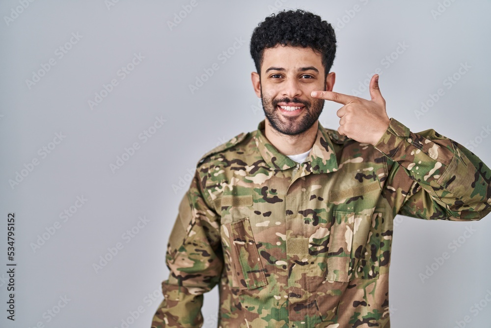 Arab man wearing camouflage army uniform pointing with hand finger to face and nose, smiling cheerful. beauty concept
