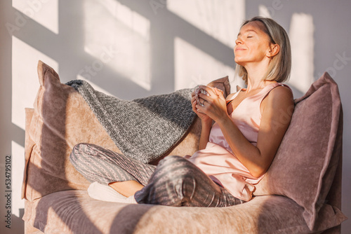 Happy dreamy middle aged woman sitting on sofa in living room with cup of tea or coffee