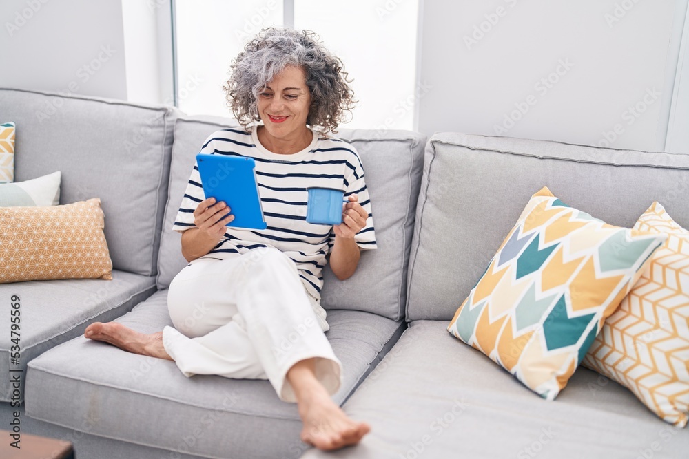 Middle age grey-haired woman using touchpad drinking coffee at home Middle age grey-haired woman using touchpad drinking coffee at home